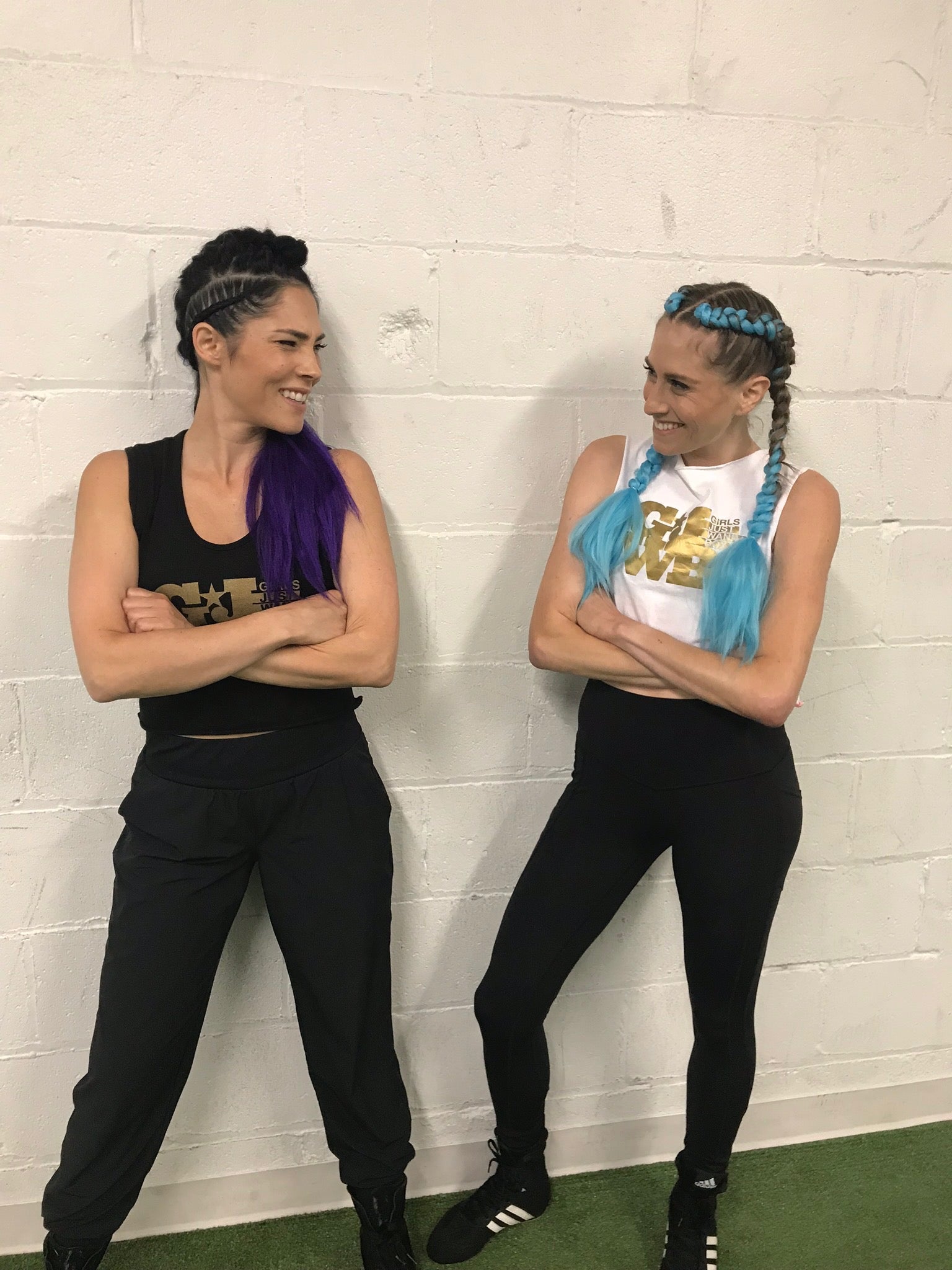 Two women dressed in workout clothes standing against a white wall with their arms crossed smiling at each other. 