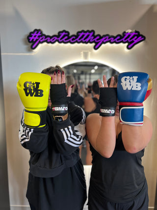 2 girls holding hands up with one hand having a black handwrap and the other with a boxing glove on 