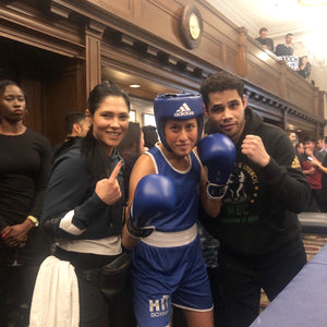 A women in black holds up her index finger signing #1. Another women in the middle in blue boxing gear is in a fighting stance. A man beside her holding up his fist dressed in black.