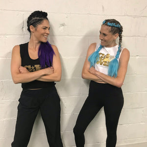 Two women dressed in workout clothes standing against a white wall with their arms crossed smiling at each other. 